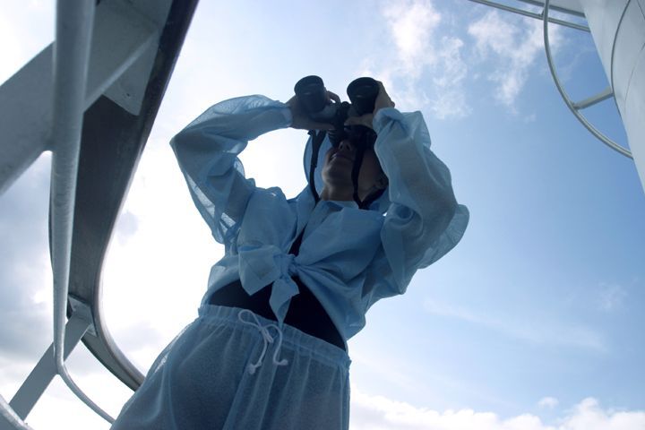 A woman wearing loose, white clothing stands on the deck of a boat looking through binoculars
