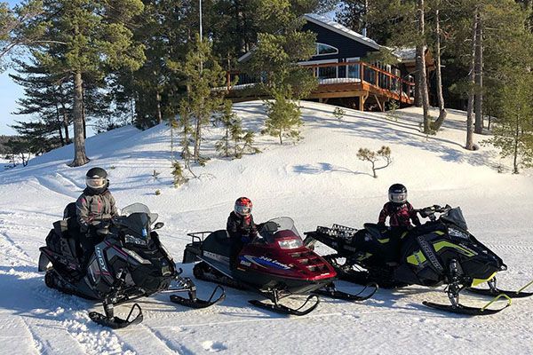 1 adult and two kids on snowmobiles in front of a cottage