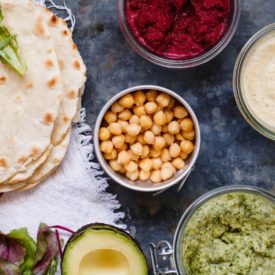 An avocado, a bowl of chickpeas, some bowls of hummus, and some pita bread laid out on a table.