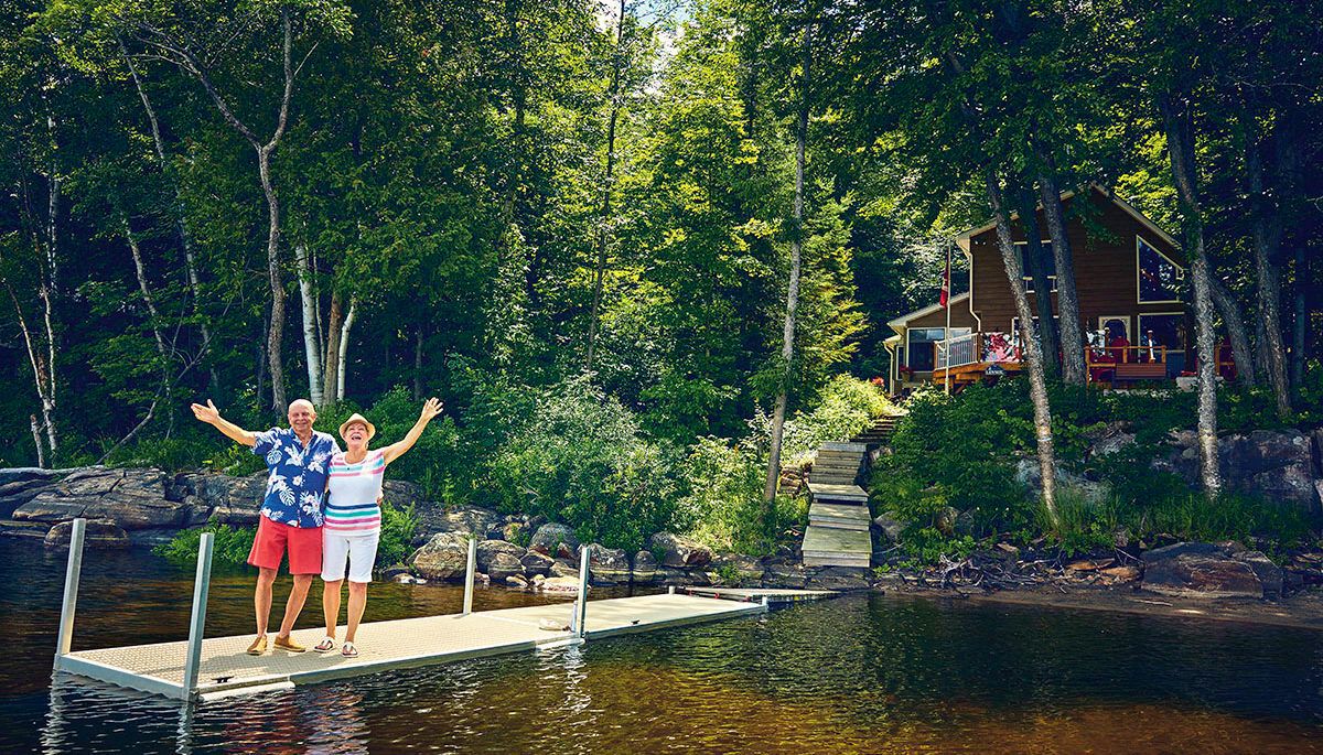 older retired couple waving goodbye from a dock in front of a cottage on a lake