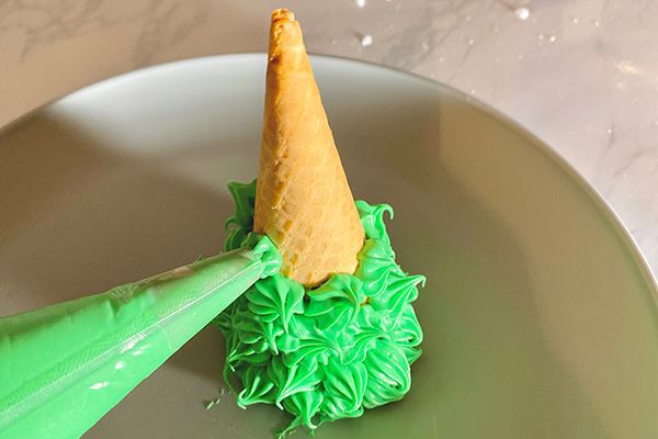 ice cream cone being covered in green frosting to make a tree for the gingerbread cottage