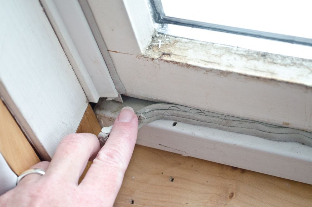 Caucasian woman's hand pressing in grey caulk in old window casement with bare wood windowsill for winterizing to keep out cold drafts and keep energy costs and heating bill down.