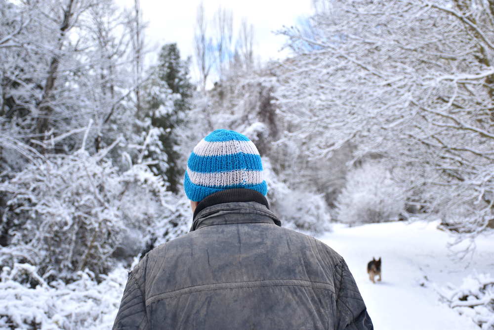 Man in old black jacket and blue and white woolen hat walking on snow-filled country road. Temporary Filomena