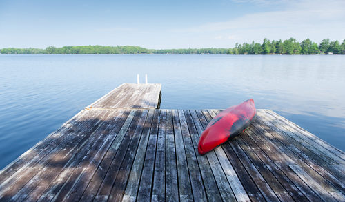 Old red kayak rest on a lake wooden dock on a summer day. Cottages are visible across the water.