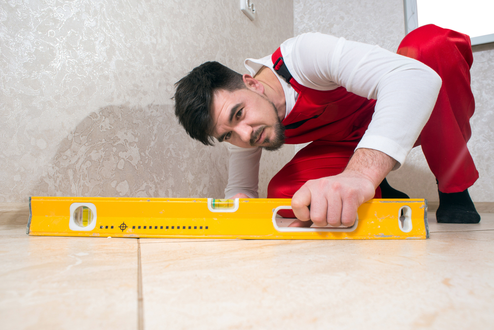 Construction worker in overalls sitting checks the quality level of flat floor tiles in the house