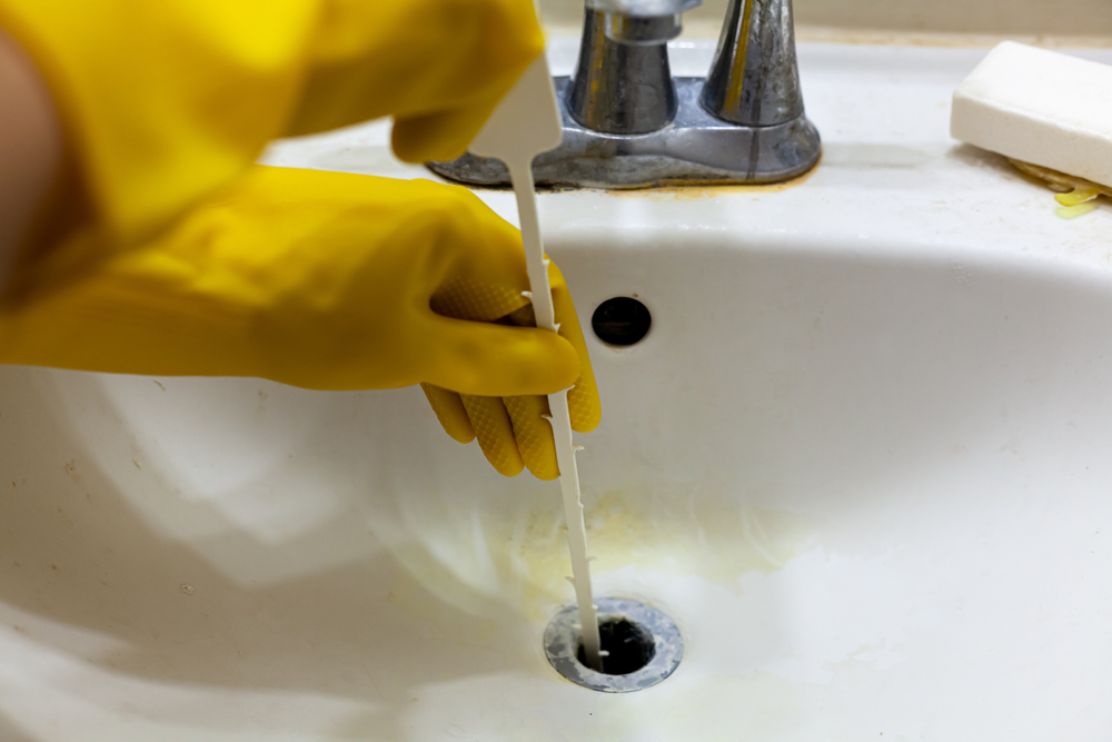 A close-up of gloved hands clearing a drain with a plastic drain tool