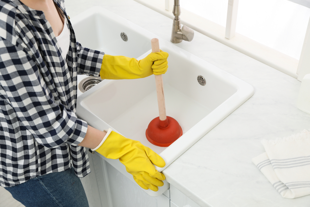 A woman with yellow rubber gloves holding a plunger in the kitchen sink