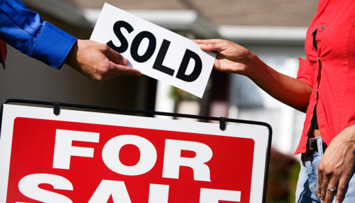 Two people exchanging a "SOLD" sign above a red-and-white For Sale sign