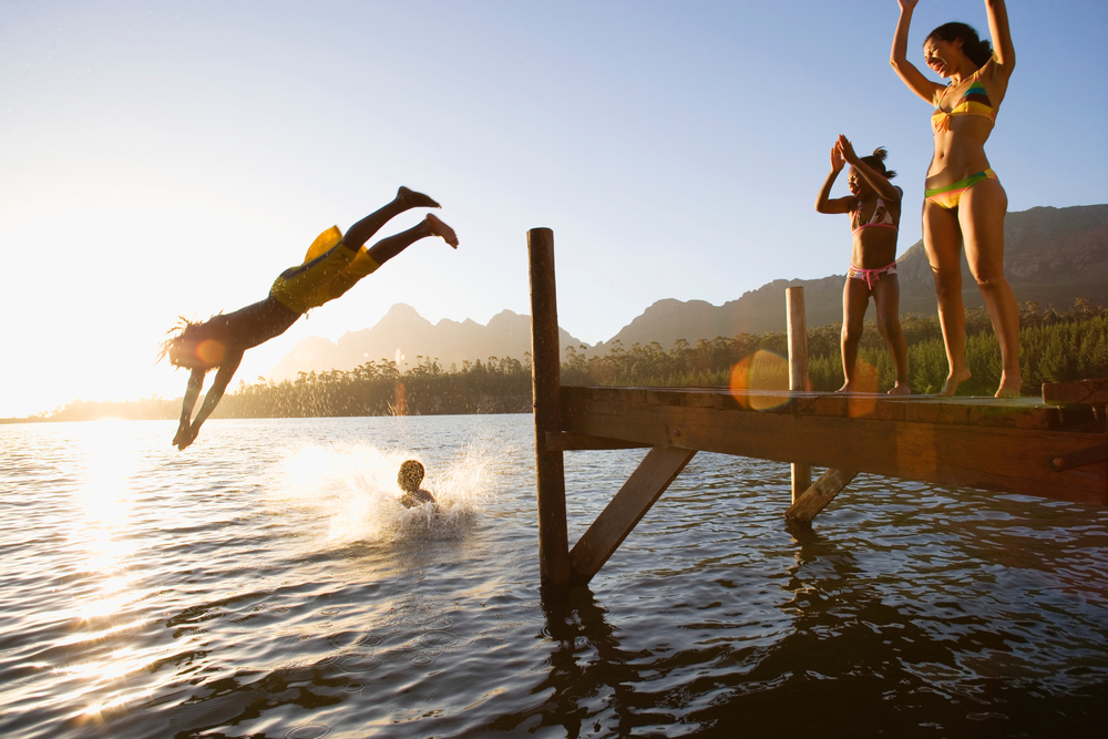 Profile shot of a father and son jumping off a dock into the lake at sunset with a splash as the daughter and mother cheers on, share a rental