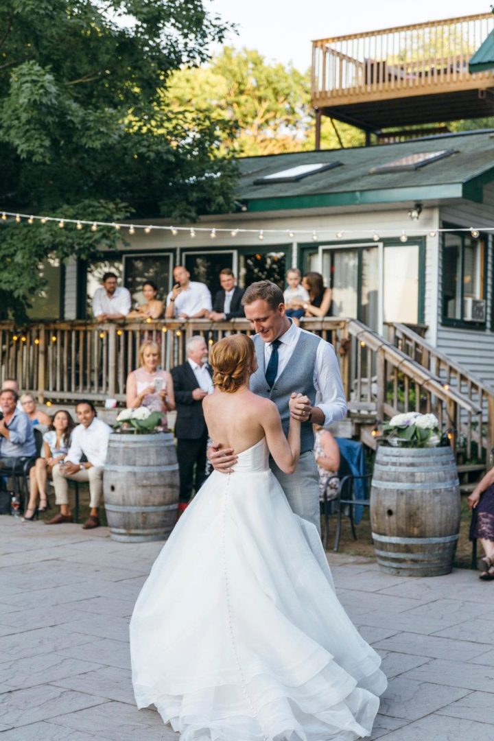 A couple dancing, dressed in wedding attire, outside in front of a cottage. Guests watching them in the background.