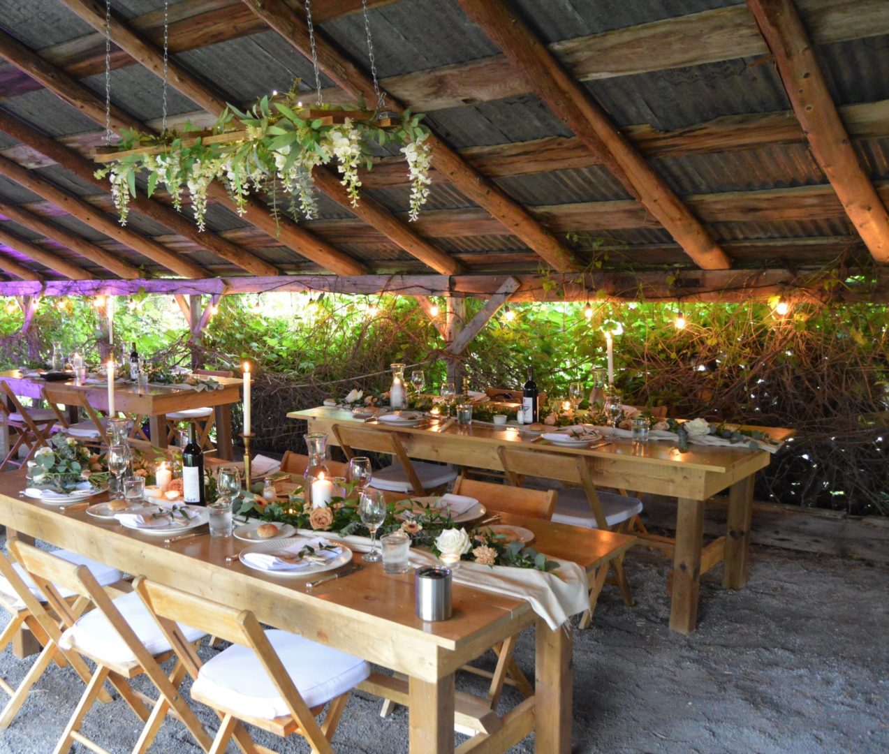 Brown wooden tables and chairs decorated with plates and linens, in a covered outdoor space.