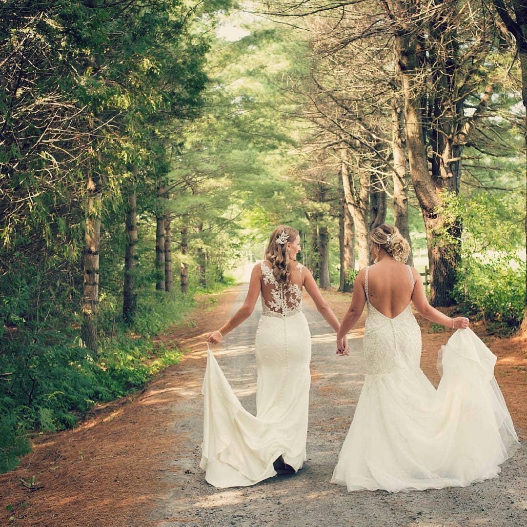 A couple in wedding gowns, holding hands and walking down a tree-lined road.