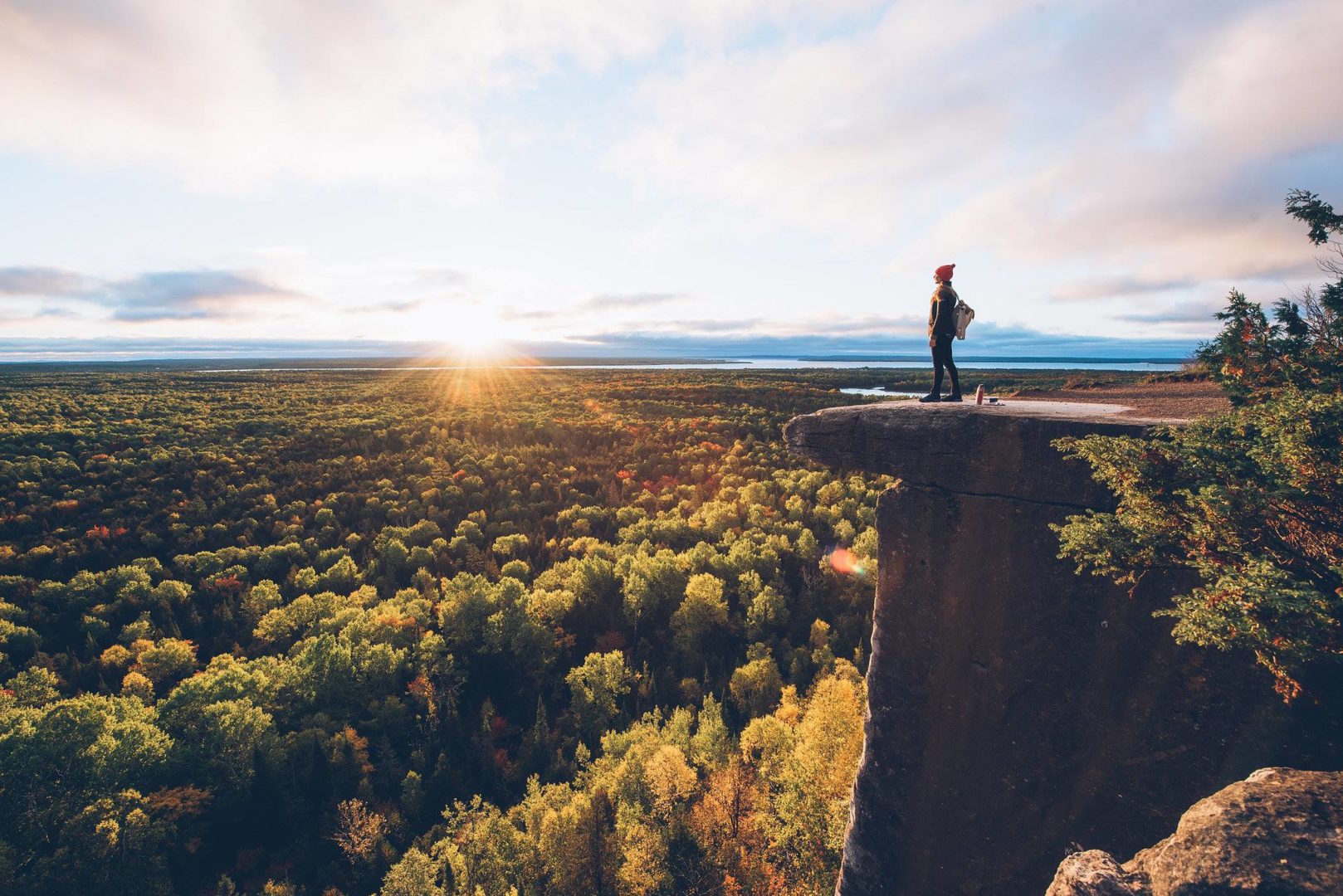 A person stands on the edge of a cliff, looking out over dense forest at sunset.
