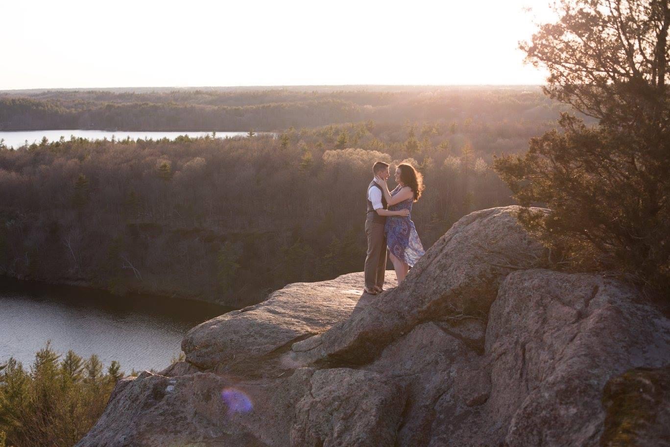 A couple stands on the edge of a rock peak, looking at each other. Lake and forest landscape in the background, at sunset.