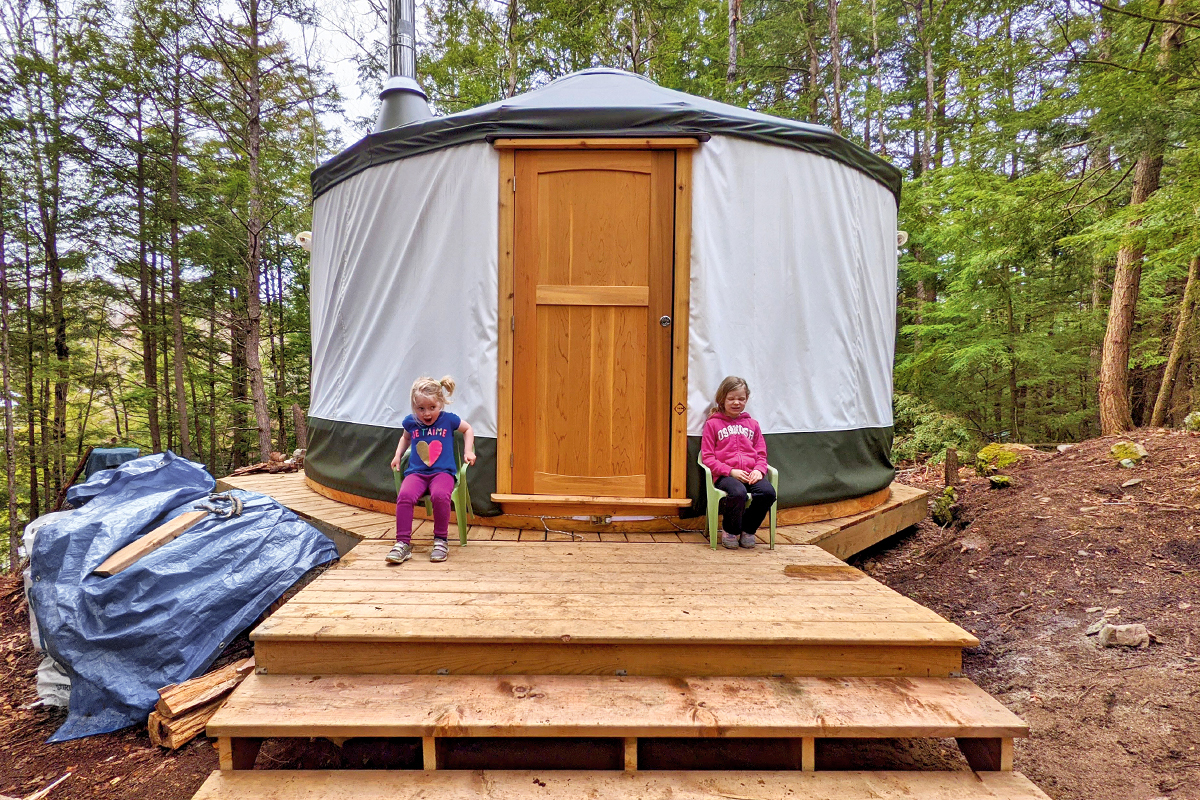 two small girls sitting outside of a yurt built on a wooden platform in a forest