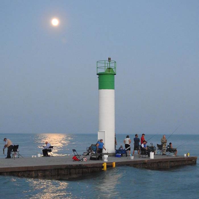 People gather on a dock and fish off the side in the early evening. A bright moon lights up the sky and the water.