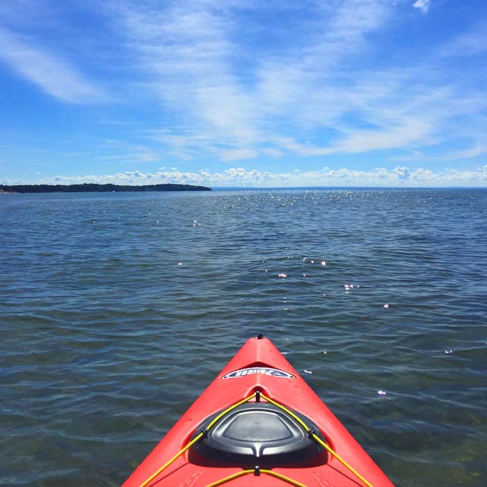 The front of a red kayak looks out onto a calm lake on a sunny day.