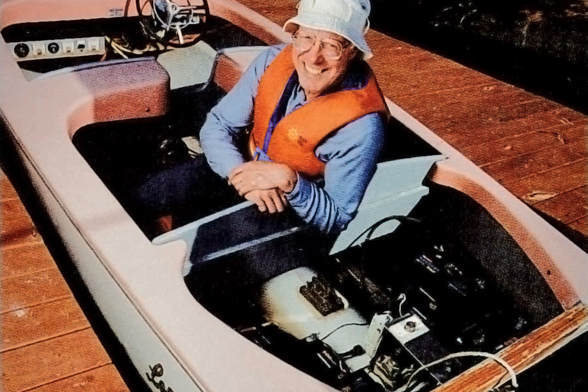 a man in a bucket hat and a life jacket sits in the solar powered wooden boat that he built
