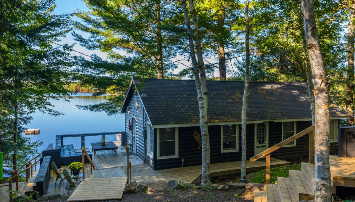 A cottage with a large deck, surrounded by tall trees with a lake in the background.