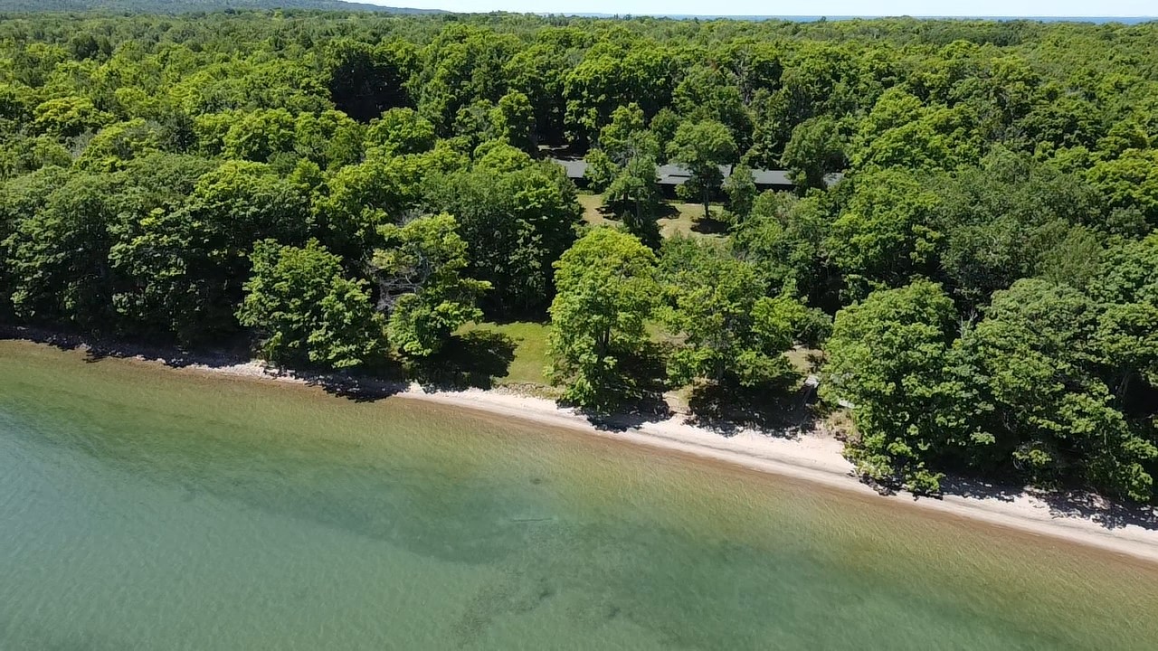 Overhead shot of a sandy stretch of shoreline on the water. A large cottage can be seen through the trees just beyond.