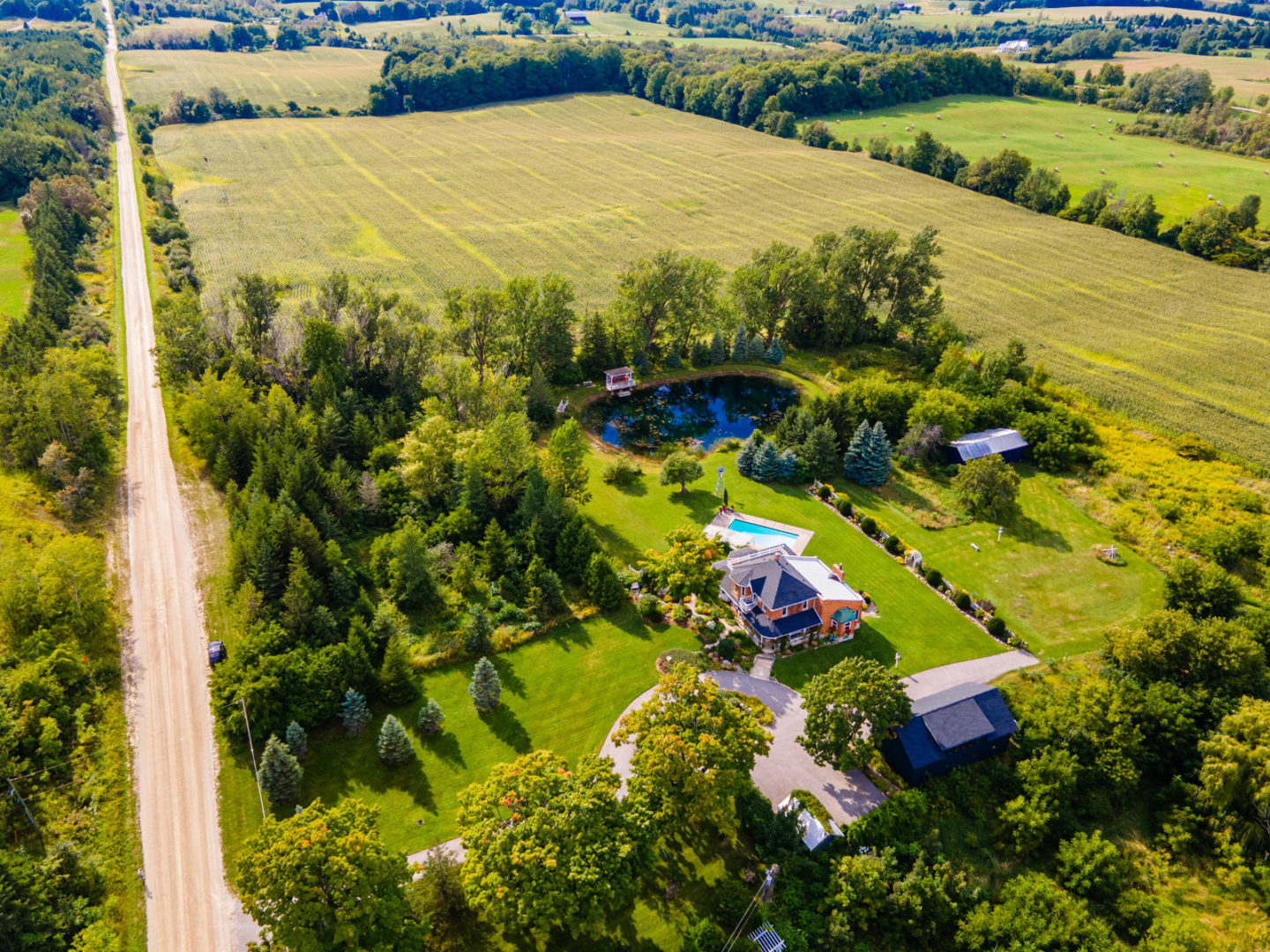 A tree-lined section of country property off a dirt road, with a large country home, a small coach house, and a private pond.