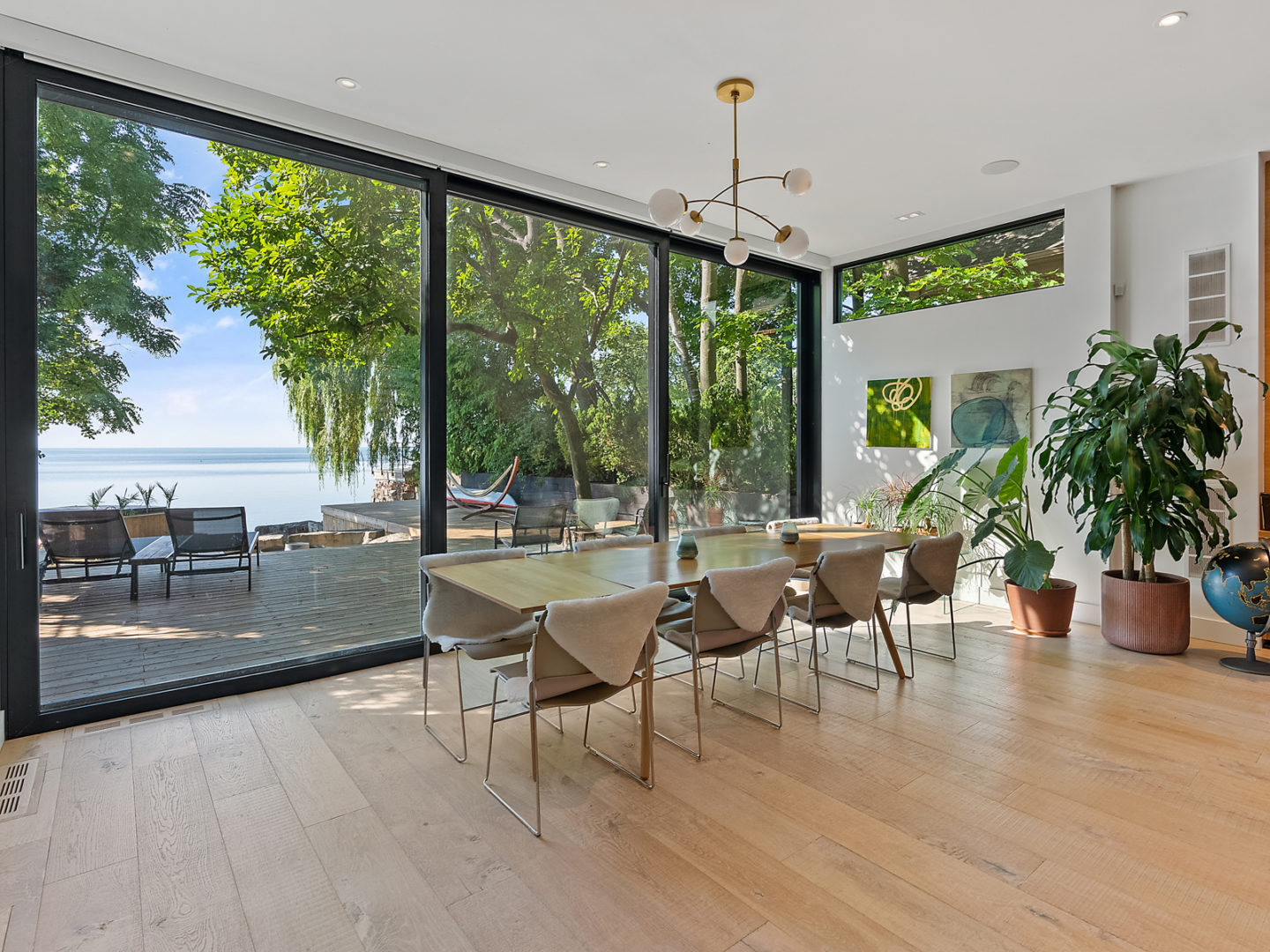 A bright dining area, with chairs and a table in from of a wall of floor-to-ceiling windows that look out on a view of a lake.