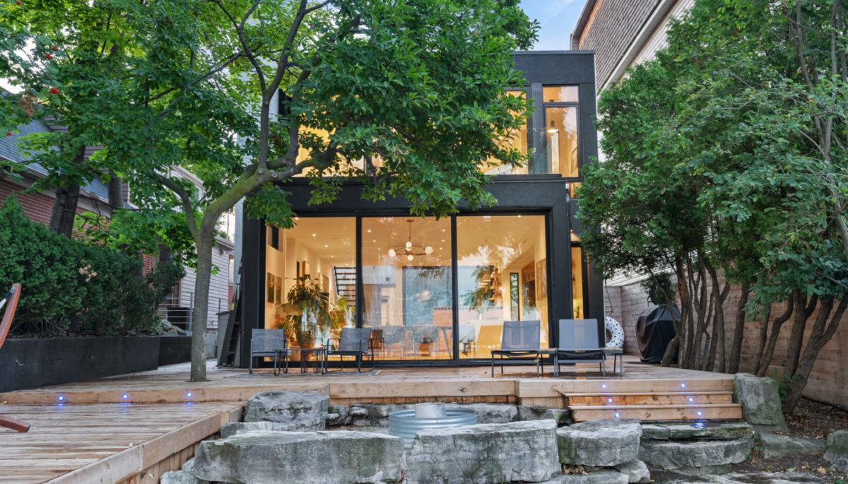 Exterior view of a modern, luxurious house with two storeys and floor-to-ceiling windows on both floors. A large deck extends outward, in front of the house.