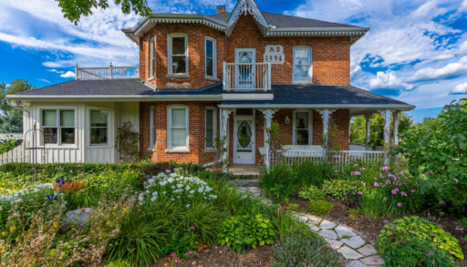 A historic country estate, two storeys high, with a large, lush garden in front of the house. The home has lots of windows, a large porch, red brick, and white trim.