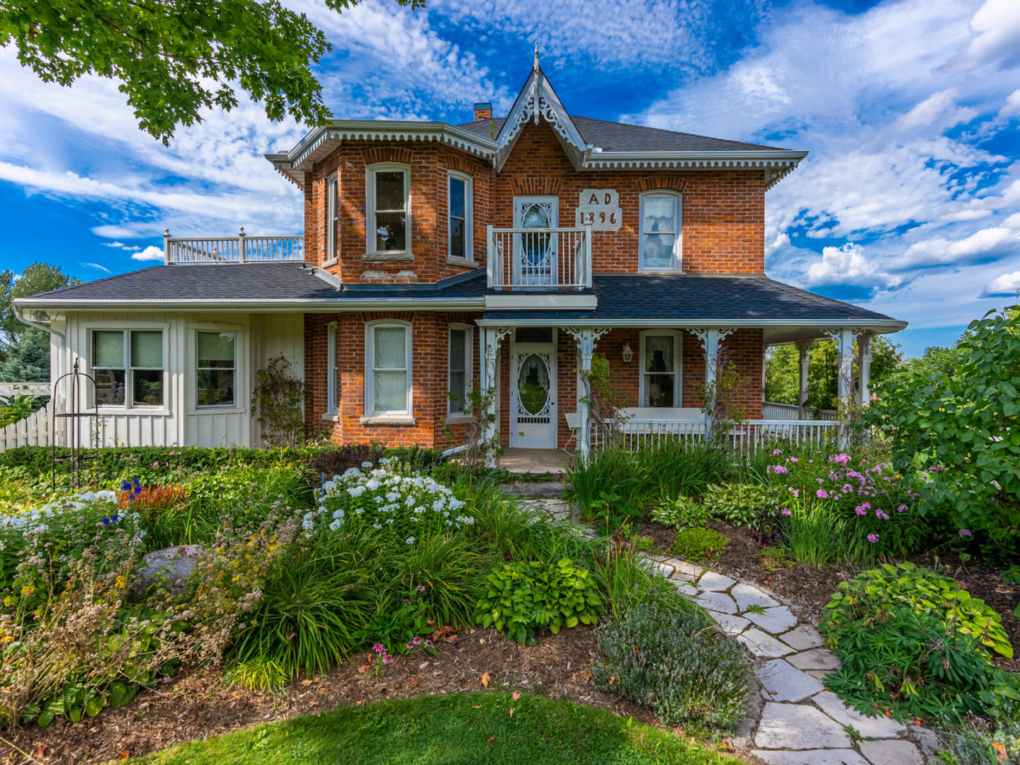 A historic country estate, two storeys high, with a large, lush garden in front of the house. The home has lots of windows, a large porch, red brick, and white trim.