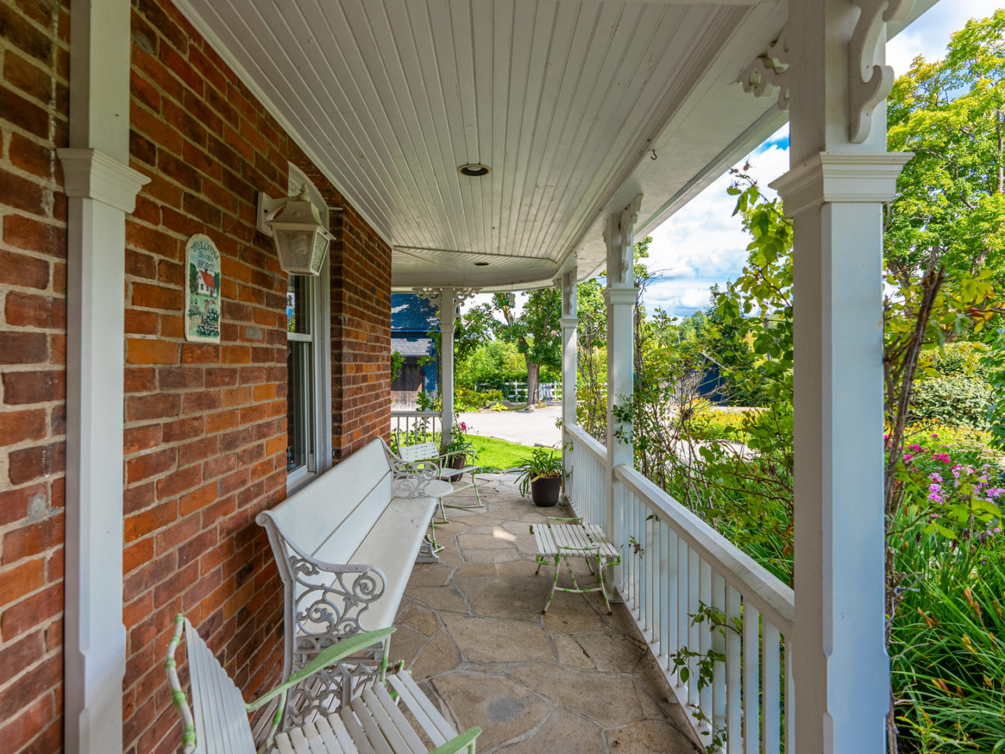 A sideways view looking down a front porch that spans the length of a red-bricked house. A white railing lines the front of the porch, and there are white benches for sitting.