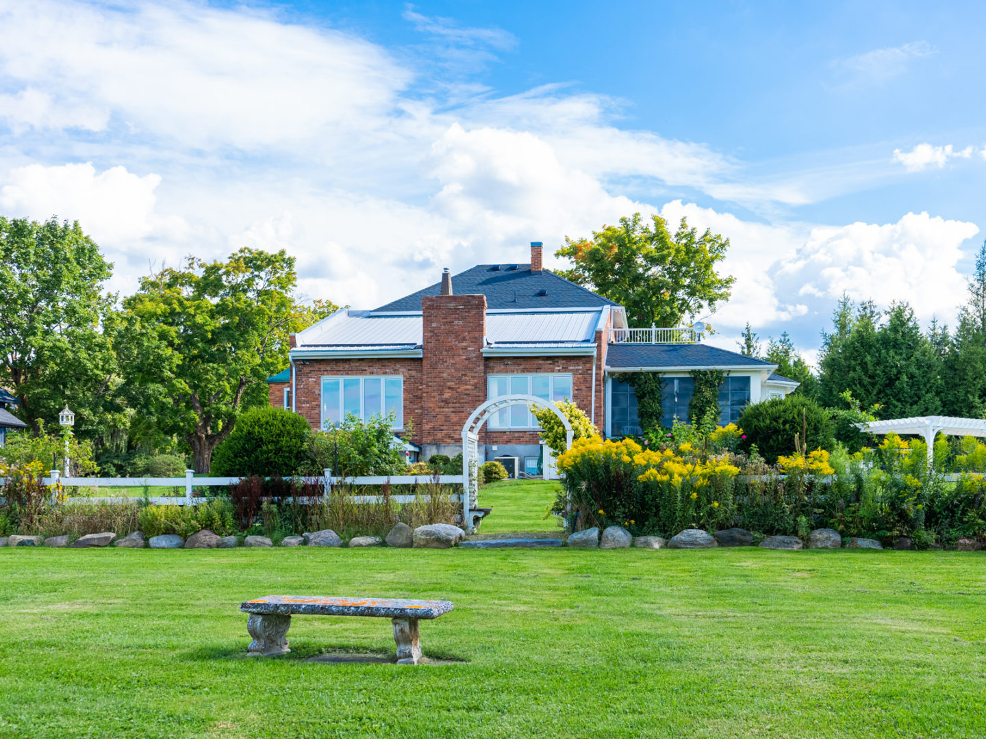A large, grassy field with a single bench in the middle. A house is in the background, past a fence with an archway.