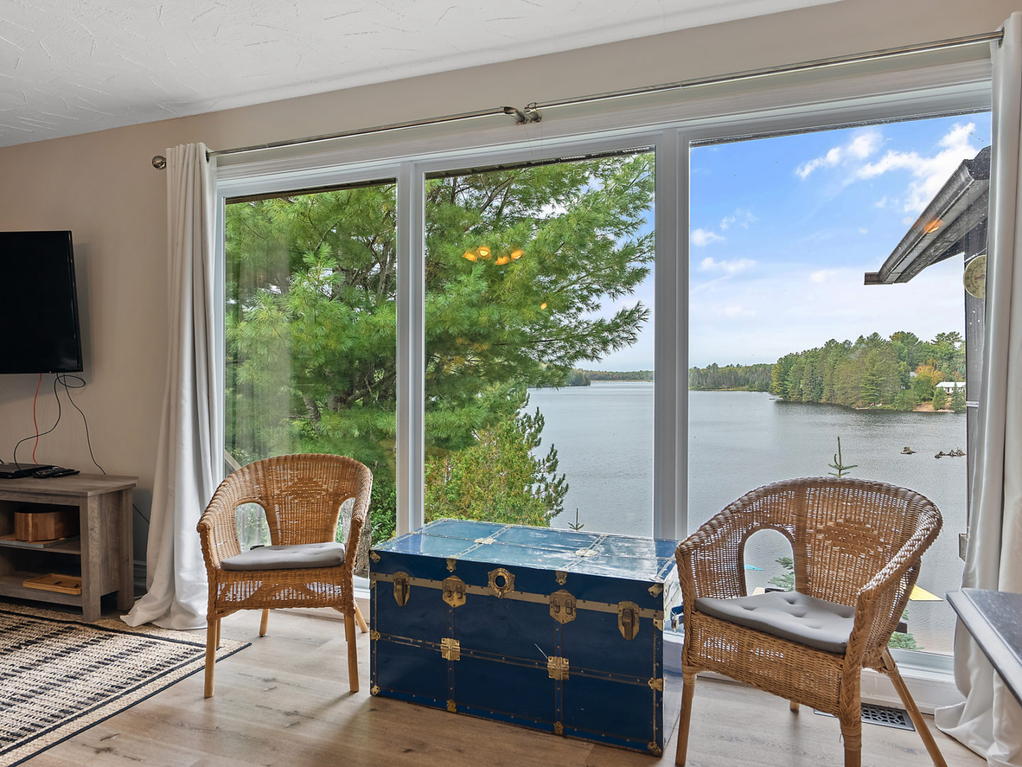 Two wicker chairs and a blue chest acting as a coffee table, in front of a large window that looks over a lake.