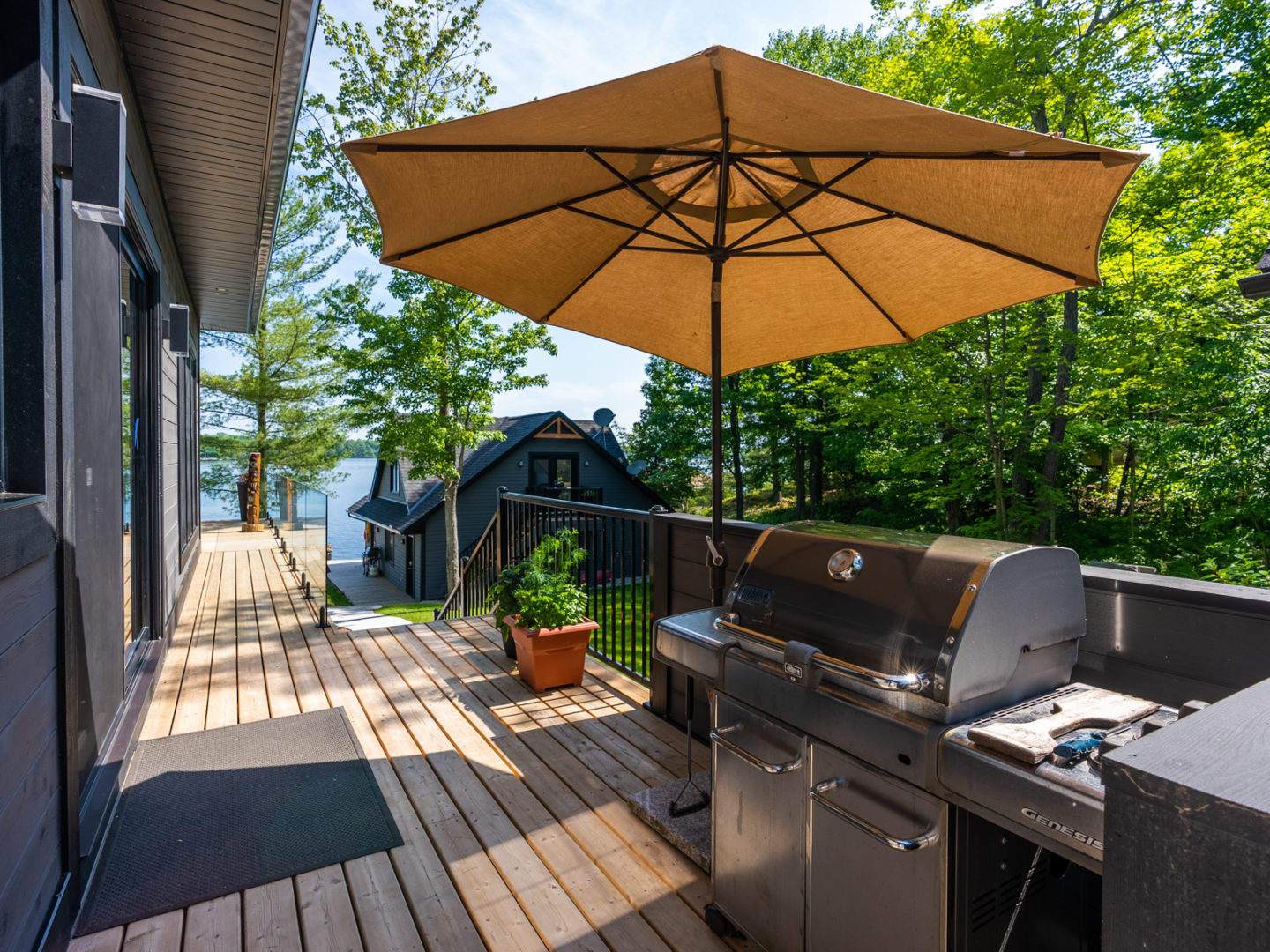 A long deck down the side of a house with a barbecue, an umbrella, and a glass railing.