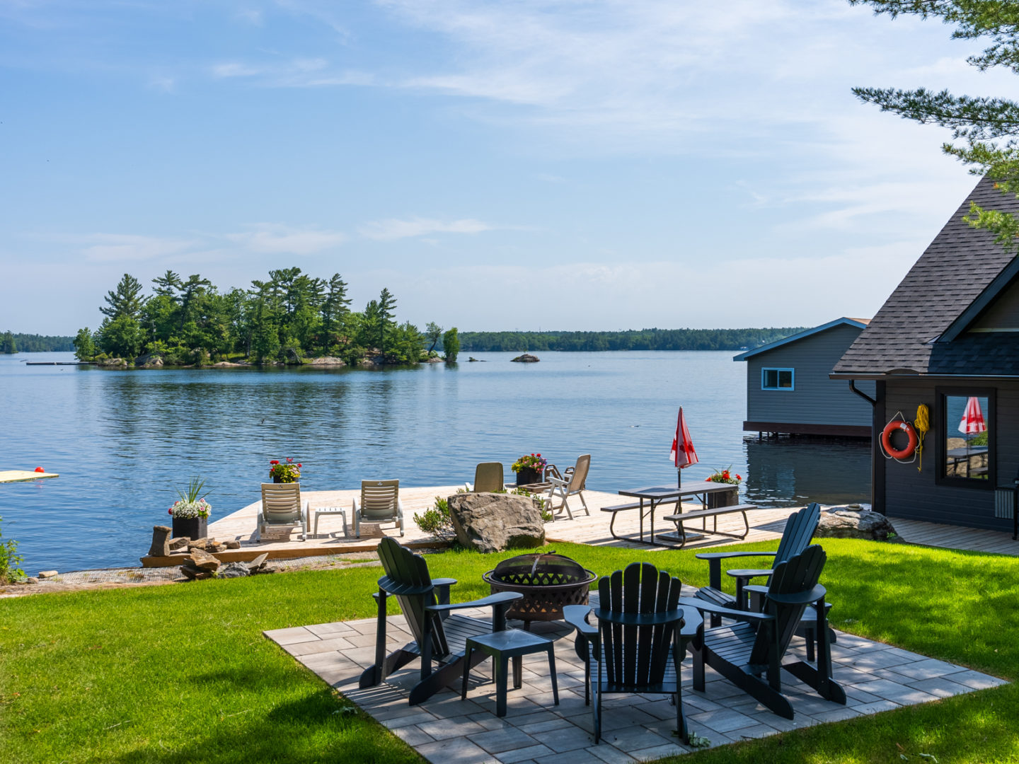 A small patio in the middle of a grassy area, with Muskoka chairs surrounding a fire pit. Down the hill is a large dock on a lake.