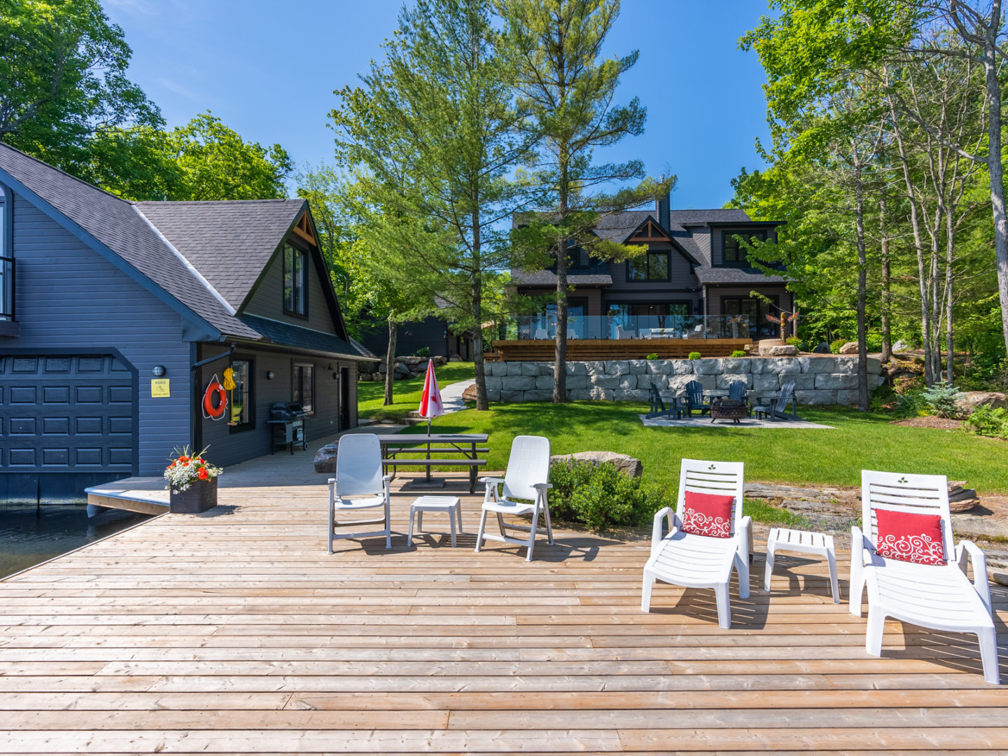 A large, wide dock in front of a boathouse, with a few dock chairs and loungers. A large cottage is in the background, up a slight hill.
