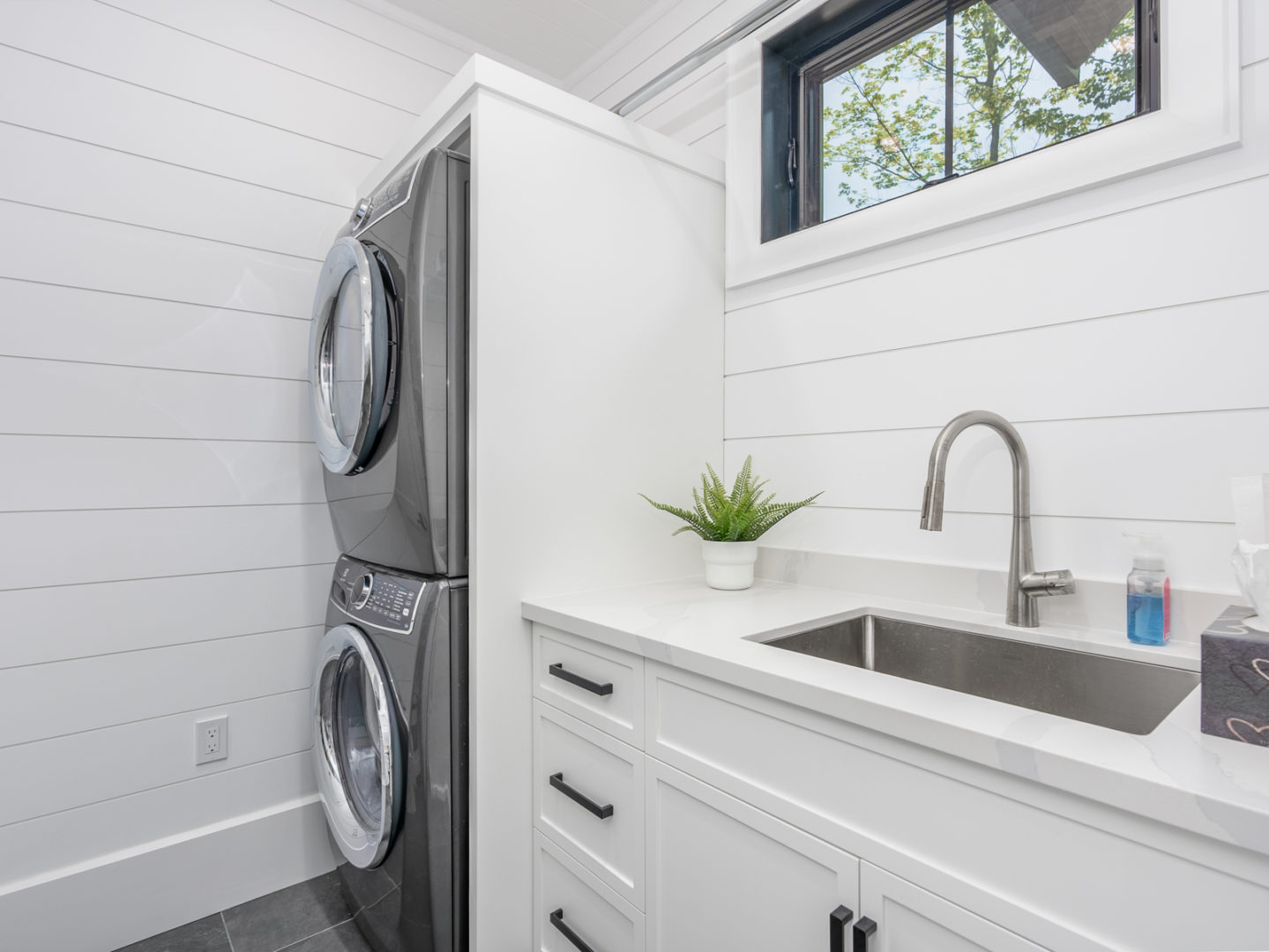 A small laundry area with a stacked washing machine and dryer, a sink area, and white walls and cupboards.