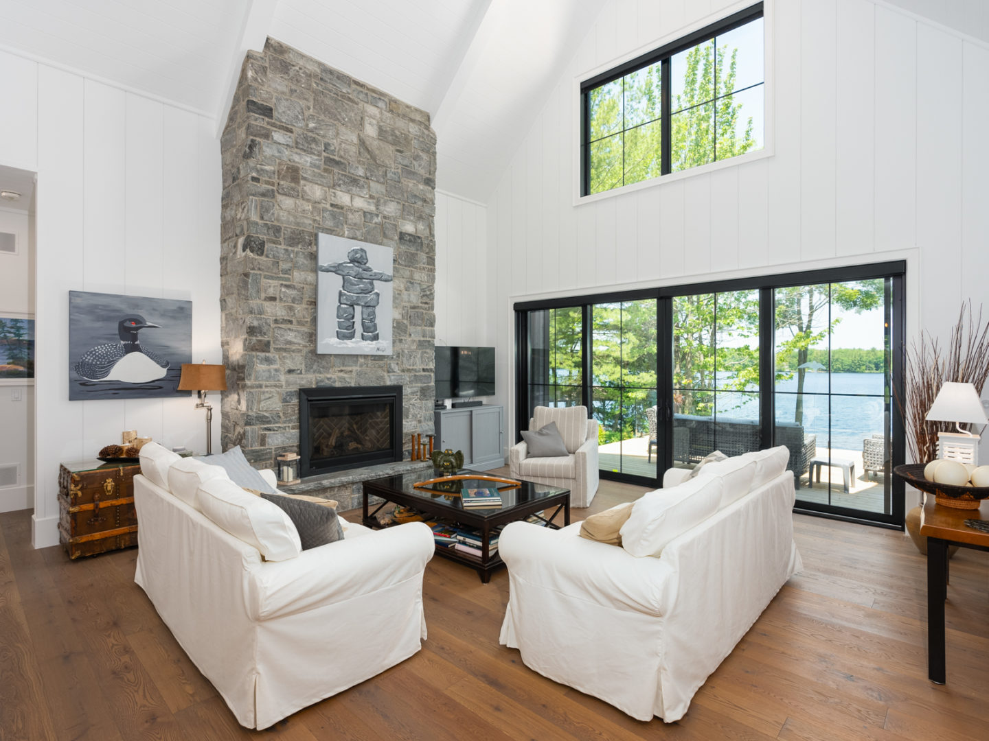 A large living space with high ceilings, wooden floors, and a stone fire place stretching two stories high. Large windows span the wall, providing a view of a lake.