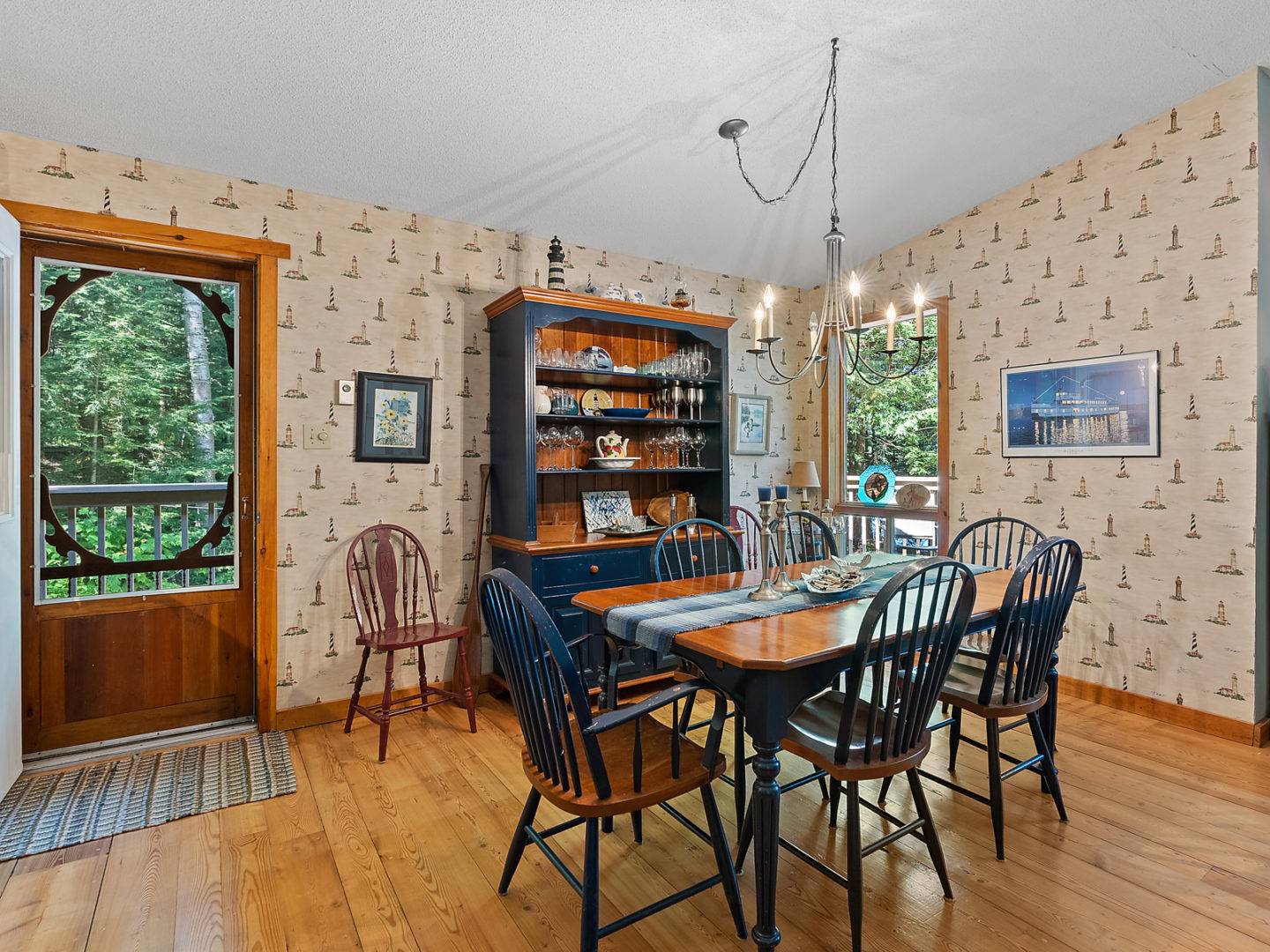 A small cottage dining area with a table and chairs. There is lighthouse wallpaper on the walls and a wooden screen door leading outside.