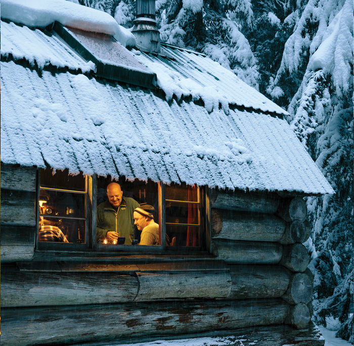 Several men gathered in a lit, snowy cabin