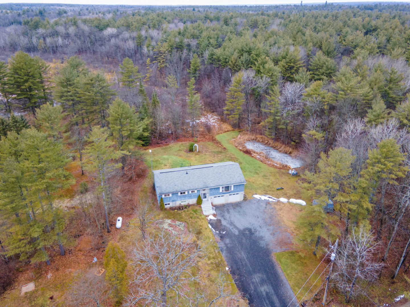 Overhead view of a modest country bungalow with a long driveway, a private pond, and forest surrounding the property.