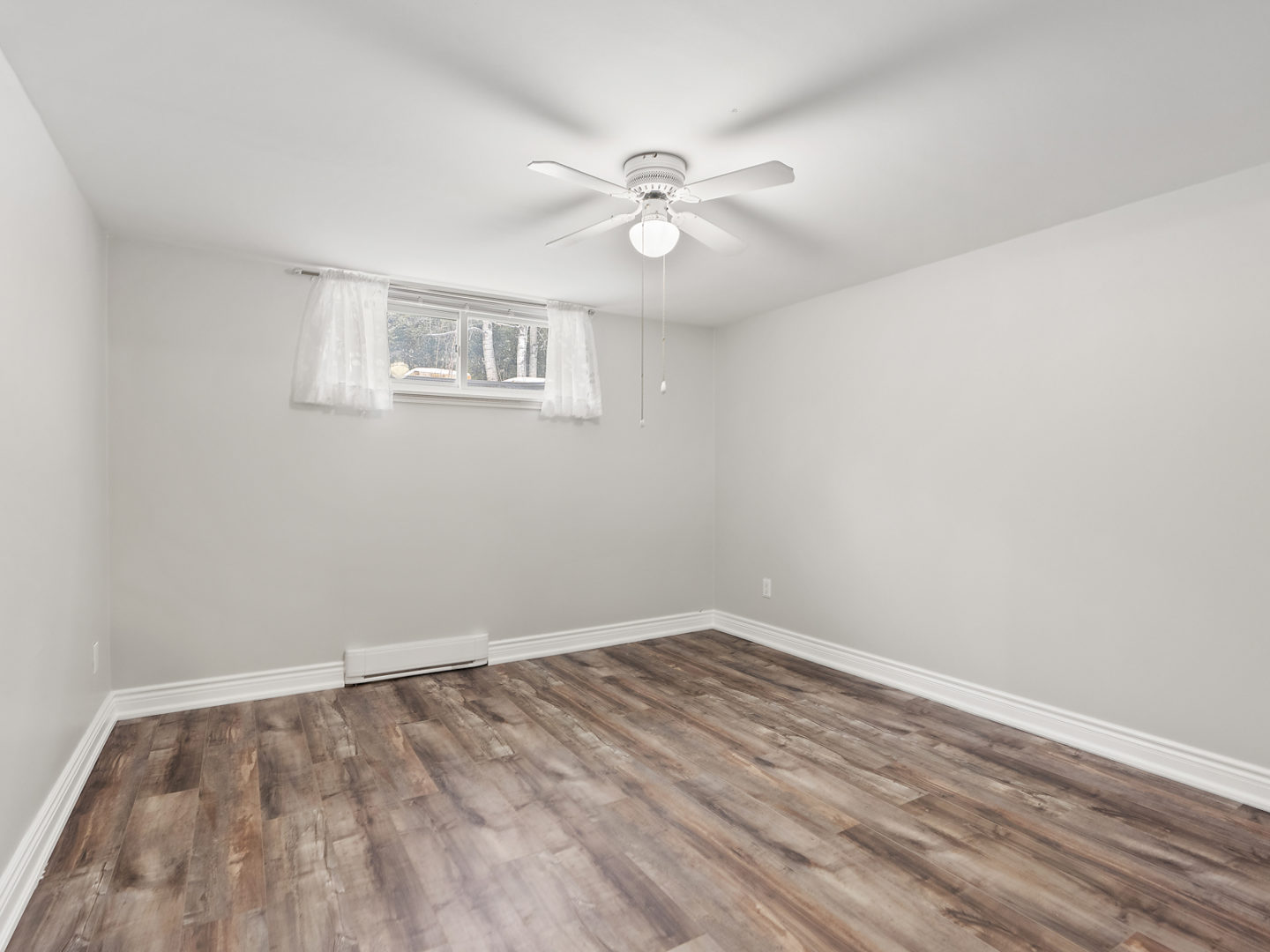 A spacious, empty room with a small window, hardwood floors, and a ceiling fan.