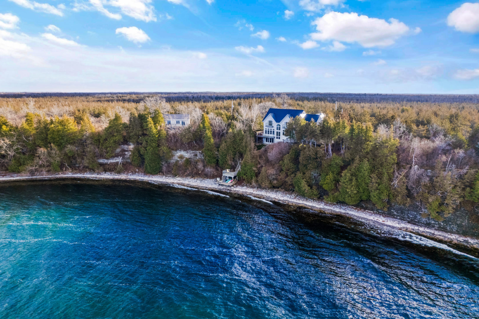 A large lake house sits on the shoreline of a deep blue lake, with a forest full of trees surrounding it.