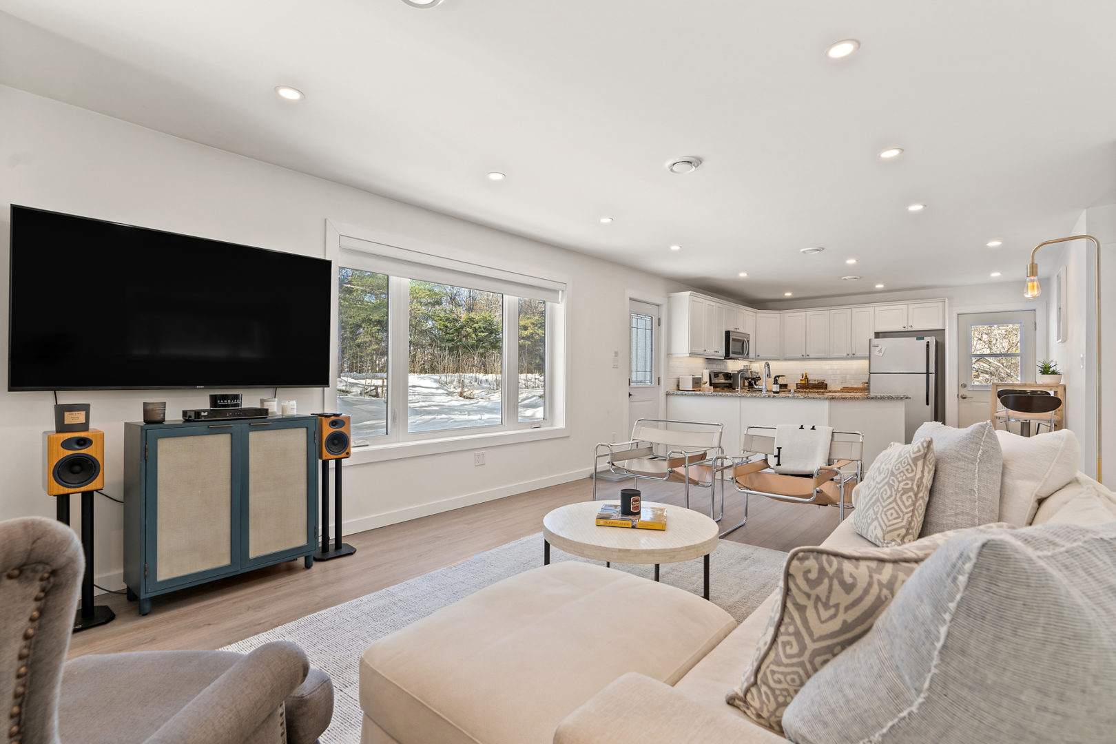 A bright living space with a couch, a television, and large windows. A kitchen is on the other side of the large open-concept space.