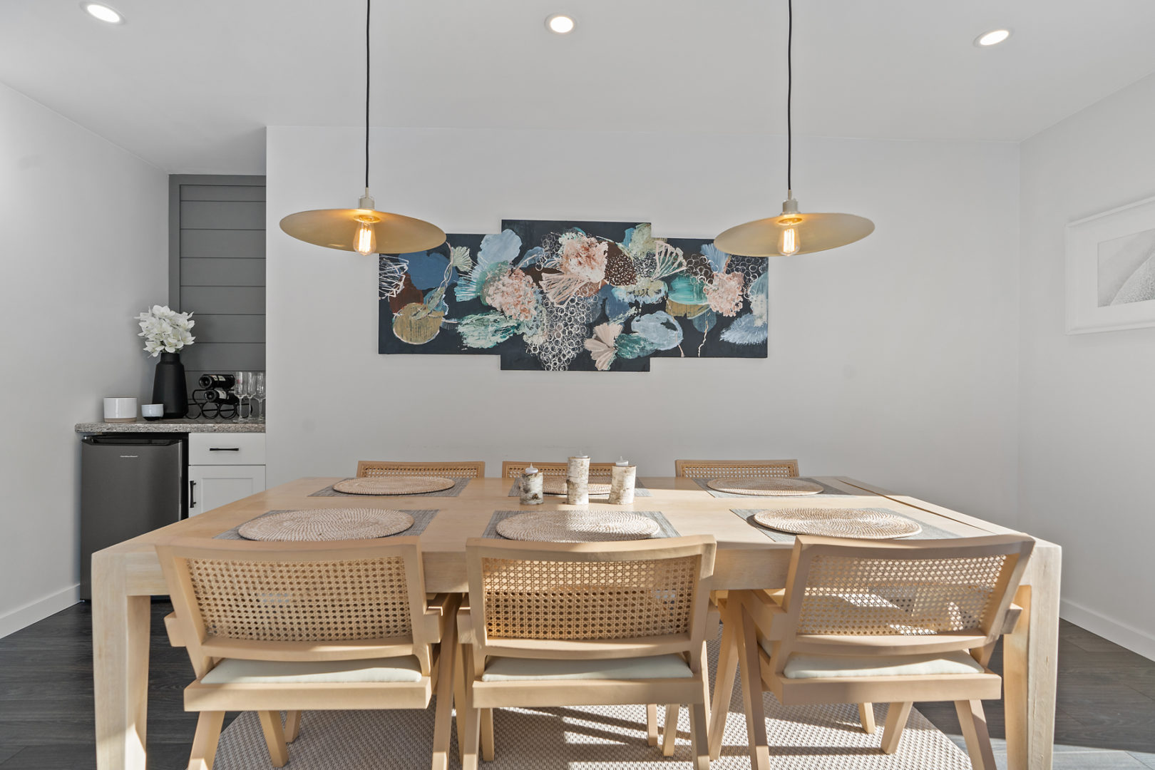 A bright dining space with a big wooden table, chairs, and overhead lighting. A small bar with a mini fridge is in the corner behind the table.