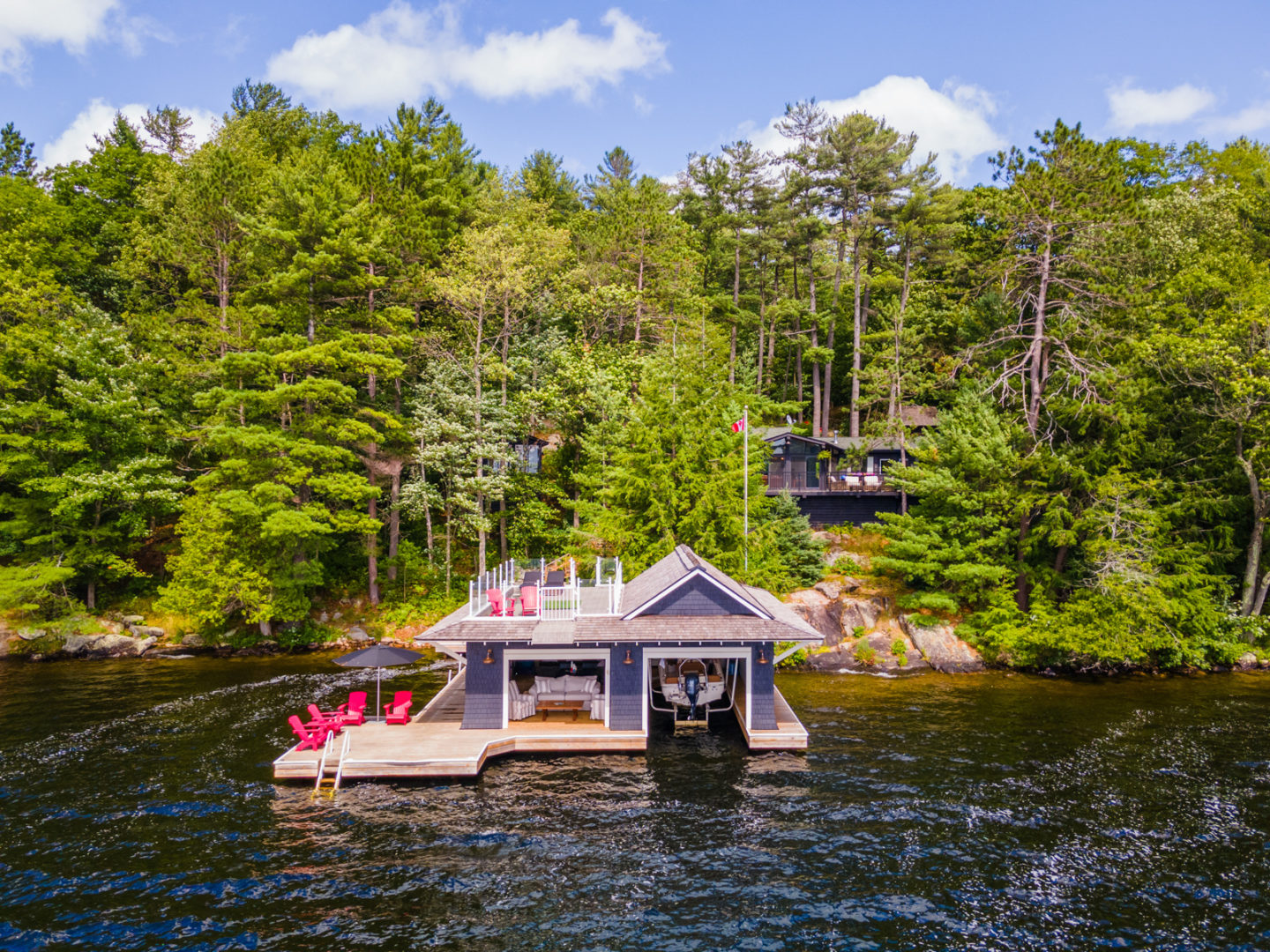 A boathouse with a large dock sits at the edge of a lake, with a cottage in behind the many trees on the shore.