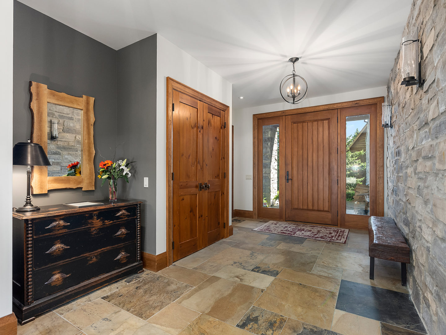 Inside the front entryway of a big house. Tiled flooring, light wood doors, and a large light fixture accent the bright space.