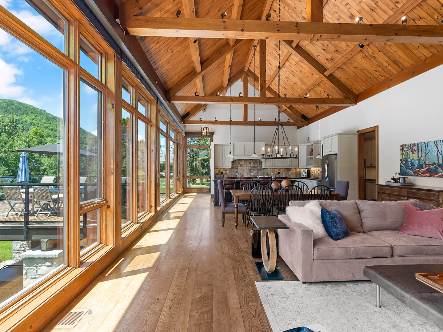 A large, open-concept living area with flow from the kitchen into the dining and living spaces. Wooden beams accent the high ceiling, and windows with a mountain view stretch all along one side of the house.