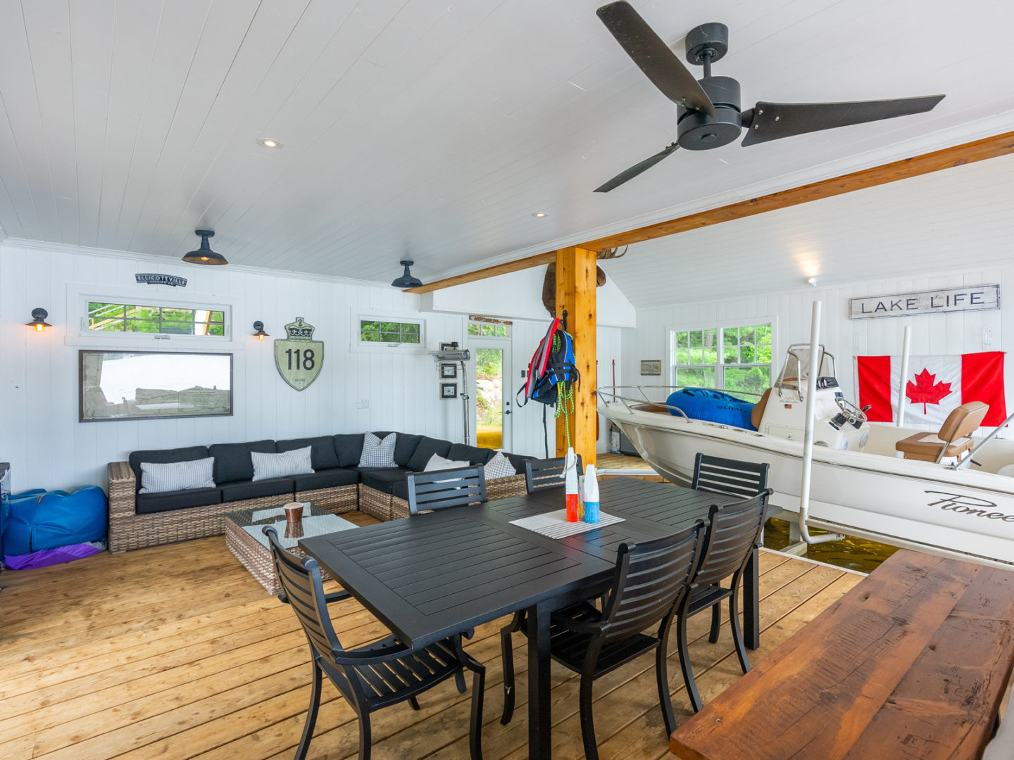 Dining area of a boathouse, with a table and chairs, a ceiling fan, a couch, and a boat parked to the side.