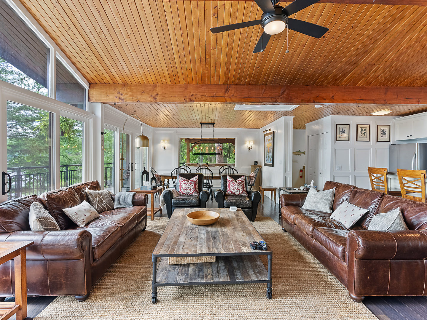 An open-concept cottage living space with large windows, wood-panelled ceilings, brown leather couches and a dining table in behind.
