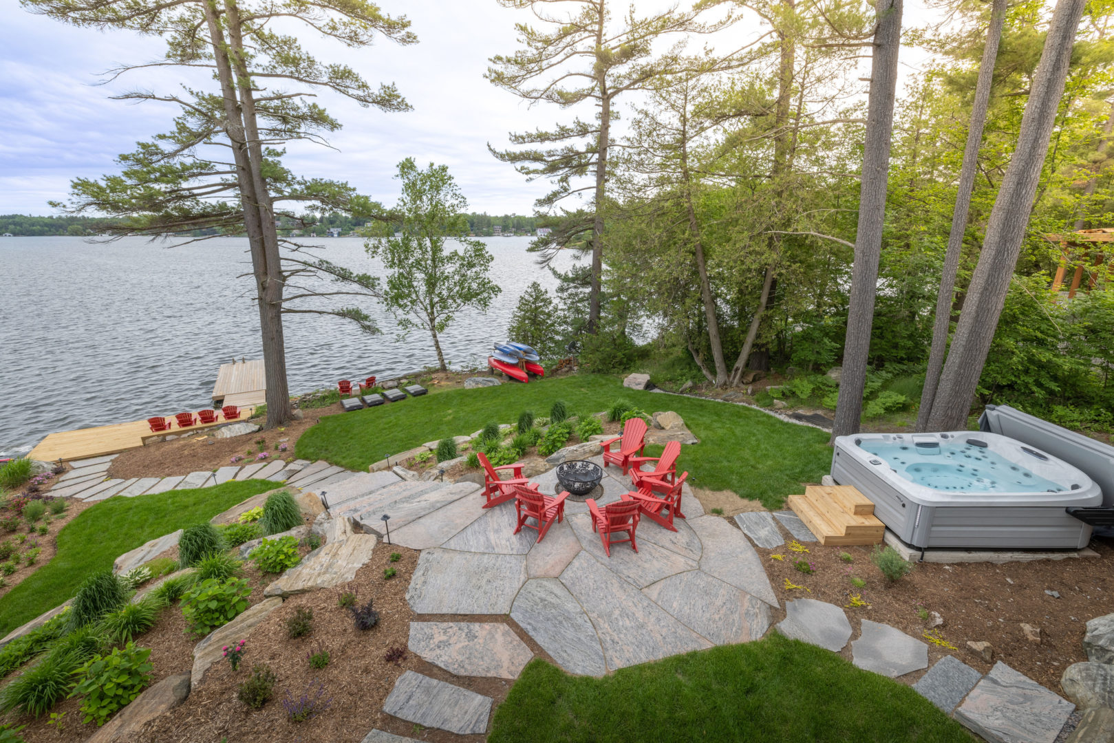 A landscaped property has a patio with a fire pit and stone steps leading down to a large lake. Trees line the property.